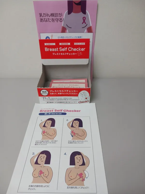 乳がん検診とセルフチェックを促すブースと、その使い方説明シートの写真。上部に「乳がん検診があなたを守る！」と書かれたピンク色のスタンドと、赤いパッケージの「Breast Self Checker（ブレストセルフチェッカー）」が箱に入っている。手前のシートには「Breast Self Checker 使い方 How to use」と題した4コマのイラストがあり、女性が片腕を上げ、反対の手で乳房全体や脇の下を順番に触ってチェックする手順が描かれている。