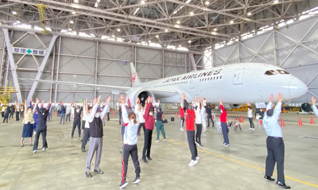 格納庫で飛行機をバックに行われているラジオ体操の様子。2人1組で向かい合って行われている