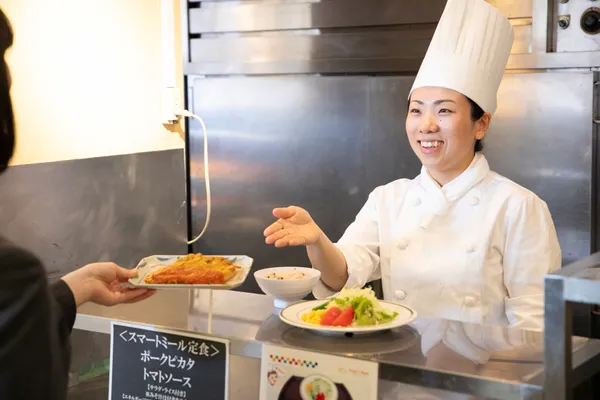 ホテル食堂のカウンターで、調理スタッフが客に料理の皿を手渡しており、カウンター上にはサラダやご飯の皿も並んでいる様子が写っている。