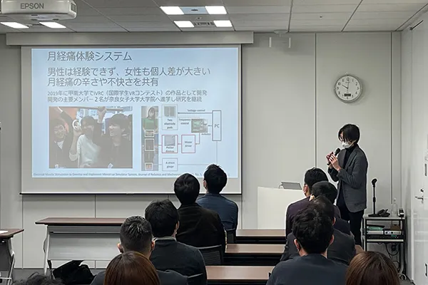 スライドを投影している講師と長机に座った受講者たち。スライドには「月経痛体験システム」と書かれている